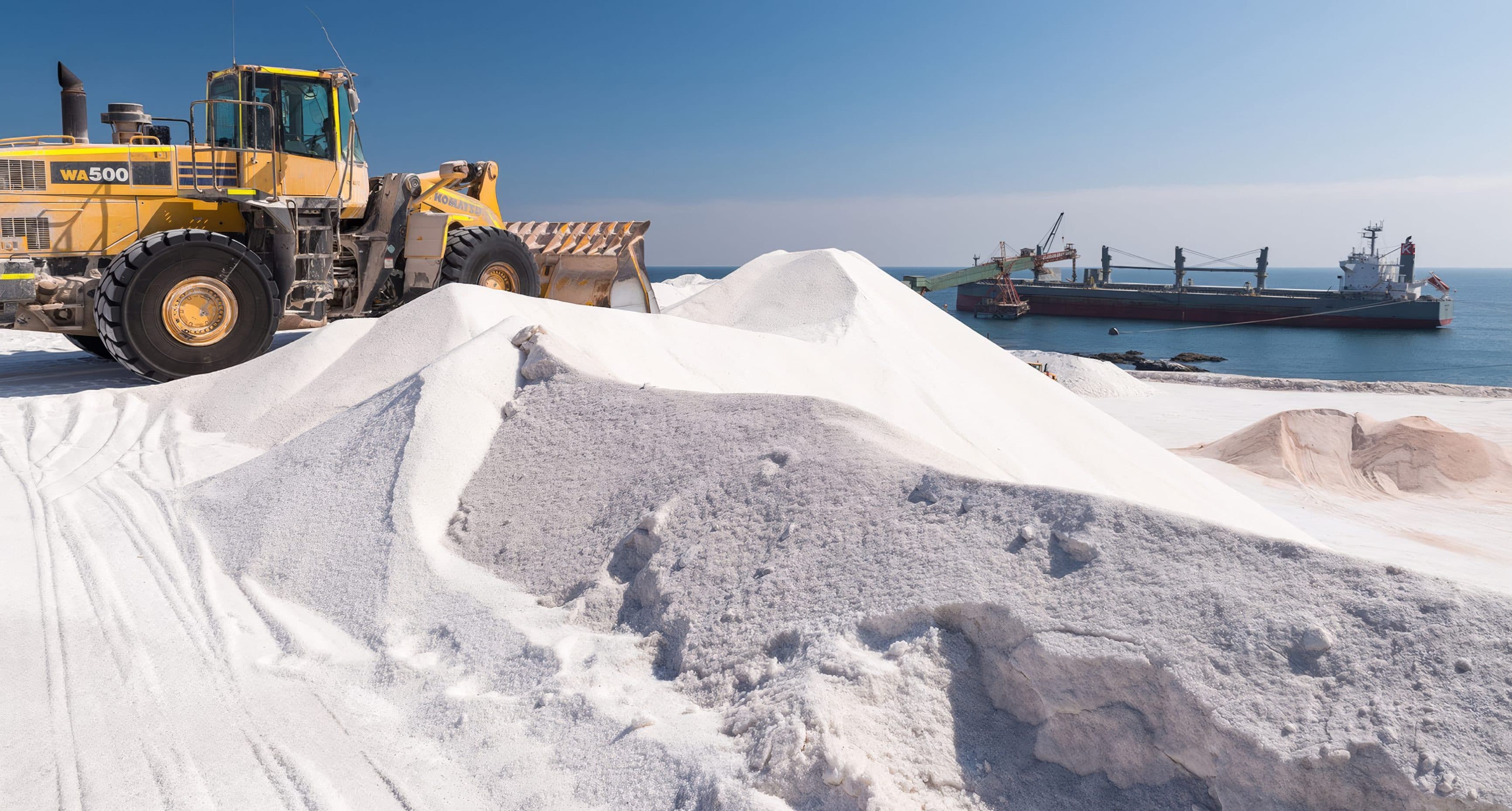 Wheel loader working salt stockpiles at a coastal port with a bulk carrier offshore